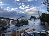 A thrilling scene at an amusement park with a roller coaster and a water ride. The water ride is creating a large splash as it descends, and the roller coaster tracks intertwine in the background. The sky is partly cloudy, and there are barrels and ropes near the water, enhancing the adventurous atmosphere.