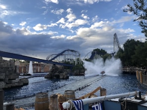 A thrilling scene at an amusement park with a roller coaster and a water ride. The water ride is creating a large splash as it descends, and the roller coaster tracks intertwine in the background. The sky is partly cloudy, and there are barrels and ropes near the water, enhancing the adventurous atmosphere.