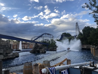 A thrilling scene at an amusement park with a roller coaster and a water ride. The water ride is creating a large splash as it descends, and the roller coaster tracks intertwine in the background. The sky is partly cloudy, and there are barrels and ropes near the water, enhancing the adventurous atmosphere.