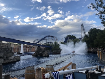 A thrilling scene at an amusement park with a roller coaster and a water ride. The water ride is creating a large splash as it descends, and the roller coaster tracks intertwine in the background. The sky is partly cloudy, and there are barrels and ropes near the water, enhancing the adventurous atmosphere.