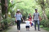 A group of people walking along a path surrounded by lush greenery. Two adults are holding the hands of a small child wearing a hat. Another person in the background appears to be watching over another child.