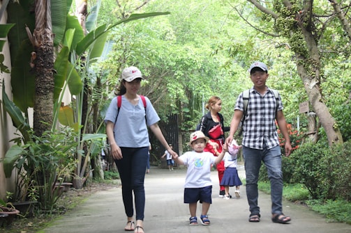 A group of people walking along a path surrounded by lush greenery. Two adults are holding the hands of a small child wearing a hat. Another person in the background appears to be watching over another child.
