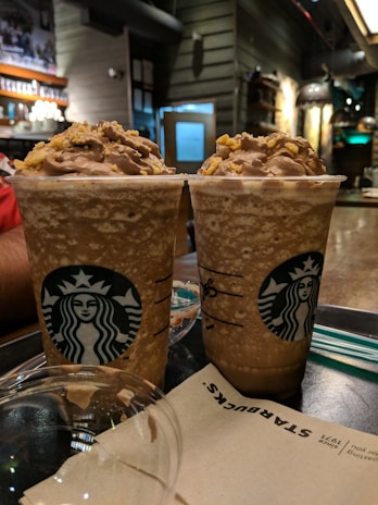 Two cold coffee beverages topped with whipped cream and caramel sprinkles are placed on a table in a cozy café setting. The interior features warm lighting, wooden accents, and several shelves with merchandise. A napkin with branding is in the foreground.