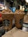 Two cold coffee beverages topped with whipped cream and caramel sprinkles are placed on a table in a cozy café setting. The interior features warm lighting, wooden accents, and several shelves with merchandise. A napkin with branding is in the foreground.