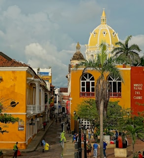 A vibrant street scene in a historic town features colorful colonial architecture. The buildings are painted in bright yellow and orange hues, and there is a large white and yellow dome visible in the background. Lush palm trees add a tropical feel, and numerous people are walking and interacting along the cobblestone path. The Museo Naval del Caribe is prominently marked on one of the buildings.