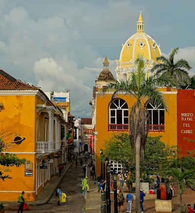 A vibrant street scene in a historic town features colorful colonial architecture. The buildings are painted in bright yellow and orange hues, and there is a large white and yellow dome visible in the background. Lush palm trees add a tropical feel, and numerous people are walking and interacting along the cobblestone path. The Museo Naval del Caribe is prominently marked on one of the buildings.