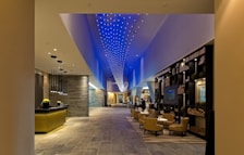 A modern, spacious hotel lobby with a contemporary design. The ceiling features a dynamic blue light installation, creating an elegant ambiance. The lobby contains a reception desk with hanging pendant lights, a lounge area with comfortable chairs and tables, and a decorative bookshelf filled with books and ornaments. The flooring appears to be made of stone or tiles, and the overall space is well-lit.