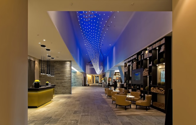 A modern, spacious hotel lobby with a contemporary design. The ceiling features a dynamic blue light installation, creating an elegant ambiance. The lobby contains a reception desk with hanging pendant lights, a lounge area with comfortable chairs and tables, and a decorative bookshelf filled with books and ornaments. The flooring appears to be made of stone or tiles, and the overall space is well-lit.