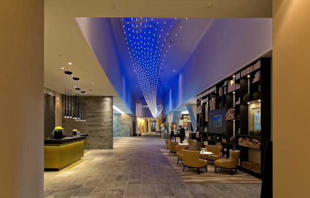 A modern, spacious hotel lobby with a contemporary design. The ceiling features a dynamic blue light installation, creating an elegant ambiance. The lobby contains a reception desk with hanging pendant lights, a lounge area with comfortable chairs and tables, and a decorative bookshelf filled with books and ornaments. The flooring appears to be made of stone or tiles, and the overall space is well-lit.
