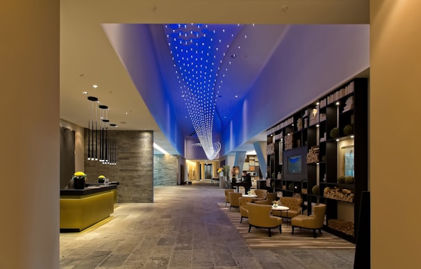A modern, spacious hotel lobby with a contemporary design. The ceiling features a dynamic blue light installation, creating an elegant ambiance. The lobby contains a reception desk with hanging pendant lights, a lounge area with comfortable chairs and tables, and a decorative bookshelf filled with books and ornaments. The flooring appears to be made of stone or tiles, and the overall space is well-lit.