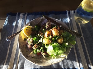A plate of food featuring a grilled fish with a lemon wedge, small roasted potatoes, and a variety of fresh greens. The dish is garnished with herbs and spices. A fork and knife are placed on the plate, and there is a glass of water with lemon on a blue and white striped tablecloth.