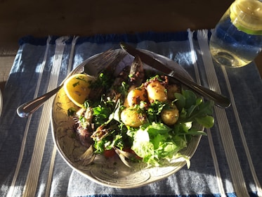 A plate of food featuring a grilled fish with a lemon wedge, small roasted potatoes, and a variety of fresh greens. The dish is garnished with herbs and spices. A fork and knife are placed on the plate, and there is a glass of water with lemon on a blue and white striped tablecloth.
