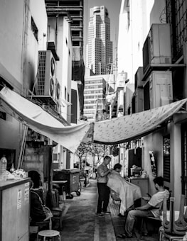 A narrow alleyway between buildings featuring a makeshift barbershop setup. A barber is cutting a customer's hair while another person sits nearby. The alley is lined with air conditioning units and is partially covered by tarps. In the background, a tall modern skyscraper rises above the older structures.