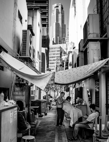 A narrow alleyway between buildings featuring a makeshift barbershop setup. A barber is cutting a customer's hair while another person sits nearby. The alley is lined with air conditioning units and is partially covered by tarps. In the background, a tall modern skyscraper rises above the older structures.