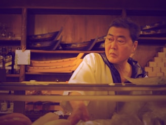 A person in a chef's uniform is standing behind a counter in what appears to be a restaurant or kitchen. Wooden shelves in the background hold empty wooden sushi boats and bottles. The chef looks focused and slightly serious.