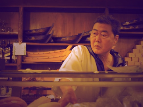 A person in a chef's uniform is standing behind a counter in what appears to be a restaurant or kitchen. Wooden shelves in the background hold empty wooden sushi boats and bottles. The chef looks focused and slightly serious.