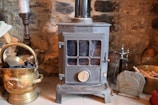 A cozy scene featuring a vintage metal stove with a glass window in the front. It is surrounded by items such as a brass coal bucket filled with coal, a wooden log, a candlestick, a small clock with Roman numerals, and a hearth brush set. The background is a rustic stone wall.