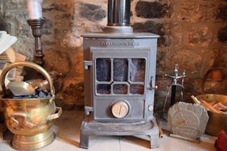 A cozy scene featuring a vintage metal stove with a glass window in the front. It is surrounded by items such as a brass coal bucket filled with coal, a wooden log, a candlestick, a small clock with Roman numerals, and a hearth brush set. The background is a rustic stone wall.