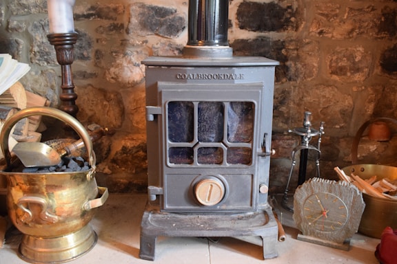 A cozy scene featuring a vintage metal stove with a glass window in the front. It is surrounded by items such as a brass coal bucket filled with coal, a wooden log, a candlestick, a small clock with Roman numerals, and a hearth brush set. The background is a rustic stone wall.