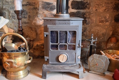 A cozy scene featuring a vintage metal stove with a glass window in the front. It is surrounded by items such as a brass coal bucket filled with coal, a wooden log, a candlestick, a small clock with Roman numerals, and a hearth brush set. The background is a rustic stone wall.
