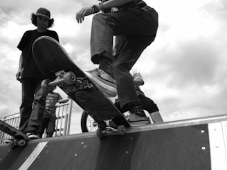 A black and white image captures a skateboarder on a ramp, displaying a low-angle perspective. The focus is on the skateboard and the rider's legs, suggesting motion and action. A couple of other individuals stand nearby, one of them holding another skateboard. The scene appears to be set outdoors, with a cloudy sky in the background.