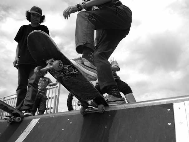 A black and white image captures a skateboarder on a ramp, displaying a low-angle perspective. The focus is on the skateboard and the rider's legs, suggesting motion and action. A couple of other individuals stand nearby, one of them holding another skateboard. The scene appears to be set outdoors, with a cloudy sky in the background.