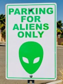 A parking sign with the text 'PARKING FOR ALIENS ONLY' in bold green letters. Below the text is a green alien head symbol with almond-shaped eyes. The sign is attached to a pole, and the background features palm trees and a clear blue sky.