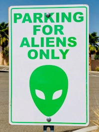 A parking sign with the text 'PARKING FOR ALIENS ONLY' in bold green letters. Below the text is a green alien head symbol with almond-shaped eyes. The sign is attached to a pole, and the background features palm trees and a clear blue sky.