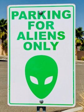 A parking sign with the text 'PARKING FOR ALIENS ONLY' in bold green letters. Below the text is a green alien head symbol with almond-shaped eyes. The sign is attached to a pole, and the background features palm trees and a clear blue sky.