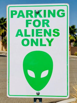 A parking sign with the text 'PARKING FOR ALIENS ONLY' in bold green letters. Below the text is a green alien head symbol with almond-shaped eyes. The sign is attached to a pole, and the background features palm trees and a clear blue sky.