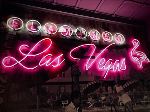 A vibrant neon sign with the words 'Flamingo Las Vegas' glowing in pink and white. The background features a vintage photograph depicting people by a pool, with umbrellas providing shade and a classic mid-century style ambiance. The image conveys a sense of nostalgia and lively atmosphere.