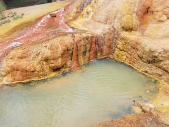 A natural hot spring with mineral-rich deposits that appear in shades of yellow, orange, and brown. Water flows gently over the rocks into a small, shallow pool. The scene is surrounded by earthy textures and colors, creating a visually diverse landscape.