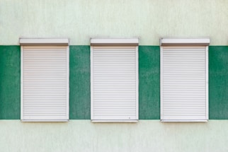 Three closed window shutters are aligned in a row on a textured wall. The wall is painted in alternating vertical green and light yellow stripes, creating a contrast between the white shutters and the background.