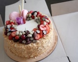 A round, layered cake topped with an assortment of fresh fruits, including strawberries, cherries, blueberries, and mulberries. Two pink macarons and an ornamental purple flower are placed on top, alongside two decorative sticks. The cake is surrounded by a crumbly, golden-brown texture.