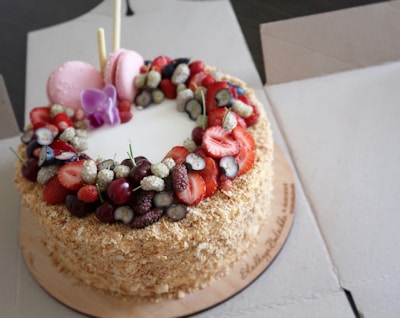 A round, layered cake topped with an assortment of fresh fruits, including strawberries, cherries, blueberries, and mulberries. Two pink macarons and an ornamental purple flower are placed on top, alongside two decorative sticks. The cake is surrounded by a crumbly, golden-brown texture.