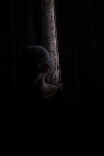 A shadowy figure lurking behind tattered curtains in a dimly lit, eerie theatre room.