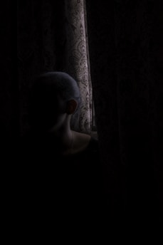 A shadowy figure lurking behind tattered curtains in a dimly lit, eerie theatre room.