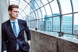 A man in a suit stands confidently by a concrete wall, under a curved metal structure overlooking a cityscape with modern buildings.