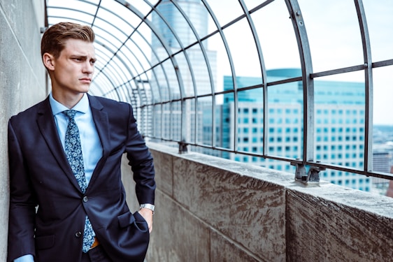 A man in a suit stands confidently by a concrete wall, under a curved metal structure overlooking a cityscape with modern buildings.