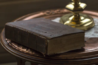 A vintage leather-bound book resting on an old wooden desk beside a softly glowing antique lamp.