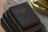 A stack of three vintage books lies on a table. The top book has a worn black cover with faint gold lettering, indicating age and frequent use. The books beneath appear similarly aged, with textures suggesting old fabric or leather binding. The background includes a blurred surface with a lace cloth and a brass object, possibly a lamp.