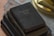 A stack of three vintage books lies on a table. The top book has a worn black cover with faint gold lettering, indicating age and frequent use. The books beneath appear similarly aged, with textures suggesting old fabric or leather binding. The background includes a blurred surface with a lace cloth and a brass object, possibly a lamp.