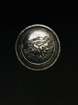 A metallic emblem with a detailed carving of a wolf's head, featuring sharp lines and intricate patterns, is centered against a dark background. The circular design includes textured borders, emphasizing the wolf motif.