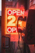 A vintage payphone is mounted on a wall next to a glass door. Behind the door, a bright neon sign is displayed with the text 'OPEN 24 OPEN' in red lights, illuminating the surroundings with a warm glow.