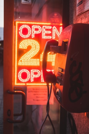 A vintage payphone is mounted on a wall next to a glass door. Behind the door, a bright neon sign is displayed with the text 'OPEN 24 OPEN' in red lights, illuminating the surroundings with a warm glow.