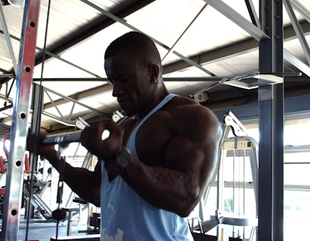 A muscular man wearing a sleeveless blue shirt is working out in a gym, performing exercises on a cable machine. The gym is spacious, with various fitness equipment visible in the background. The ceiling features metal beams and there is natural light coming through the windows.