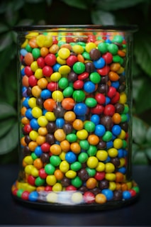 A glass jar filled with colorful, round candies featuring shades of red, green, blue, yellow, brown, and orange. The candies are densely packed, creating a vibrant and eye-catching appearance. Behind the jar, there is a blurred background of green leaves.