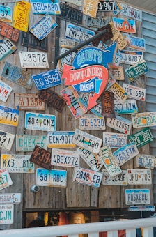 A collection of vintage license plates is displayed on a wooden wall. They come from various states and have different colors and designs. In the center, a colorful sign reads 'Geddy's Down-Under Cool Stuff' with arrows pointing in different directions. Below, part of a store with clothing items is visible.