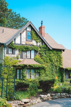 A charming inn with ivy-covered walls, large windows, and a rustic design. The building features a steep gabled roof, a central chimney, and is surrounded by a lush garden with various plants and flowers.