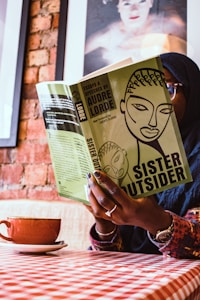 A person is sitting indoors holding a book titled 'Sister Outsider' by Audre Lorde. The cover of the book is green with an abstract illustration of a face. The person's hands are visible, displaying a ring and nail art. The background includes a brick wall and framed artwork. A cup of coffee sits on a saucer on a checkered tablecloth.